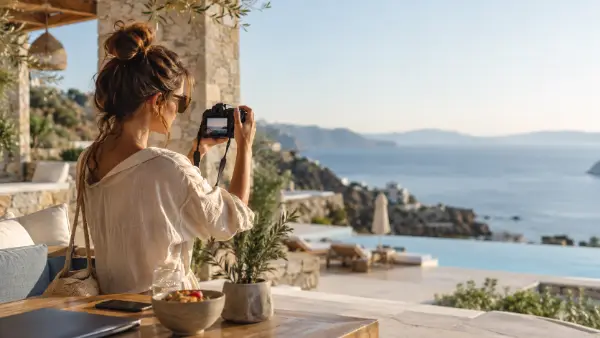 A creator with a camera on a shaded terrace above a hotel pool and the sea.
